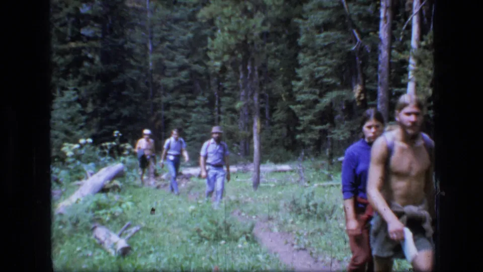 Young backpackers hike through dense Montana forest, with one shirtless man and other companions visible on the trail.