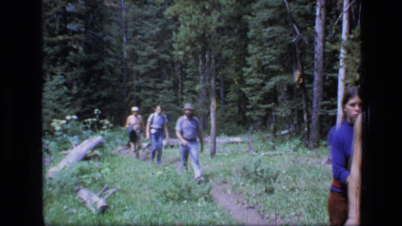 1973 8mm footage shows adult hikers approaching on a narrow trail in Scapegoat Wilderness forest, with evergreens and fallen logs around them.