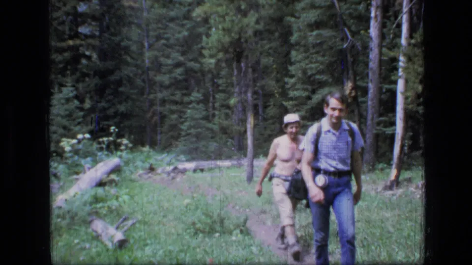 Two adult hikers approach the camera on a narrow Montana wilderness trail, with tall trees and green undergrowth behind them.