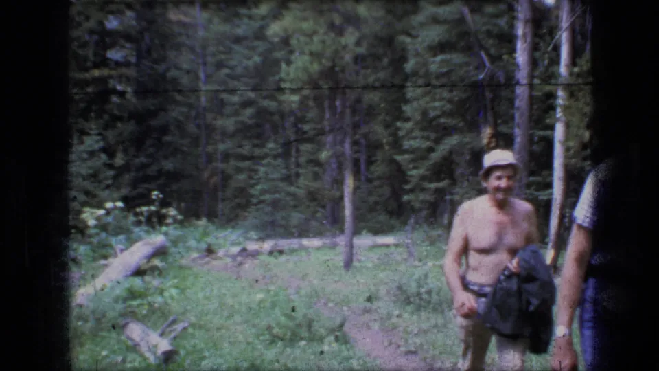 A shirtless adult hiker with a hat and carried clothing passes close to the camera on the Scapegoat Wilderness forest path.