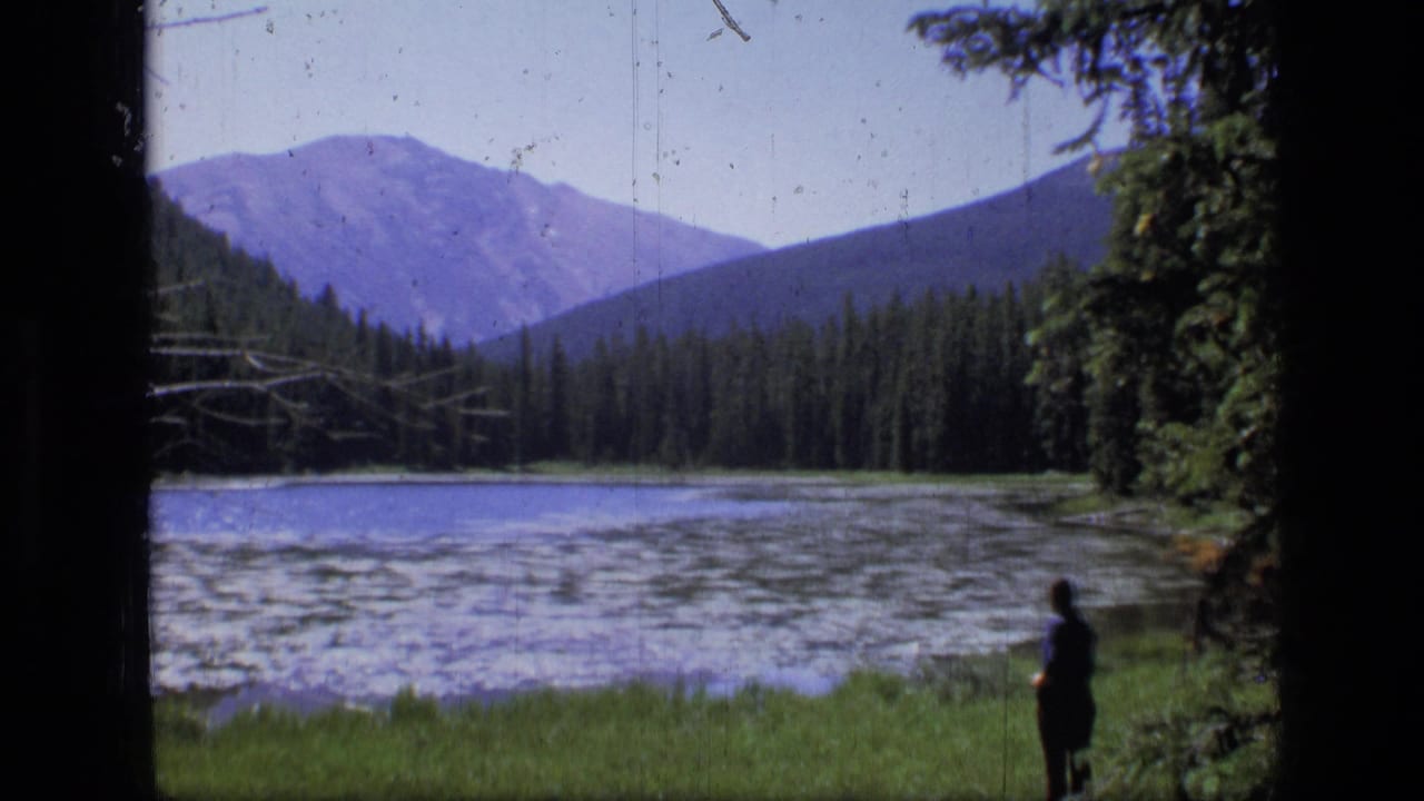 1973 8mm view of Scapegoat Wilderness, Montana, with a small figure on a grassy lakeshore below forested slopes and distant peaks.