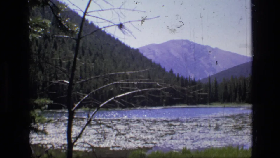 8mm home-movie frame from Scapegoat Wilderness, showing a calm lake, evergreen trees, distant mountains, and foreground branches.
