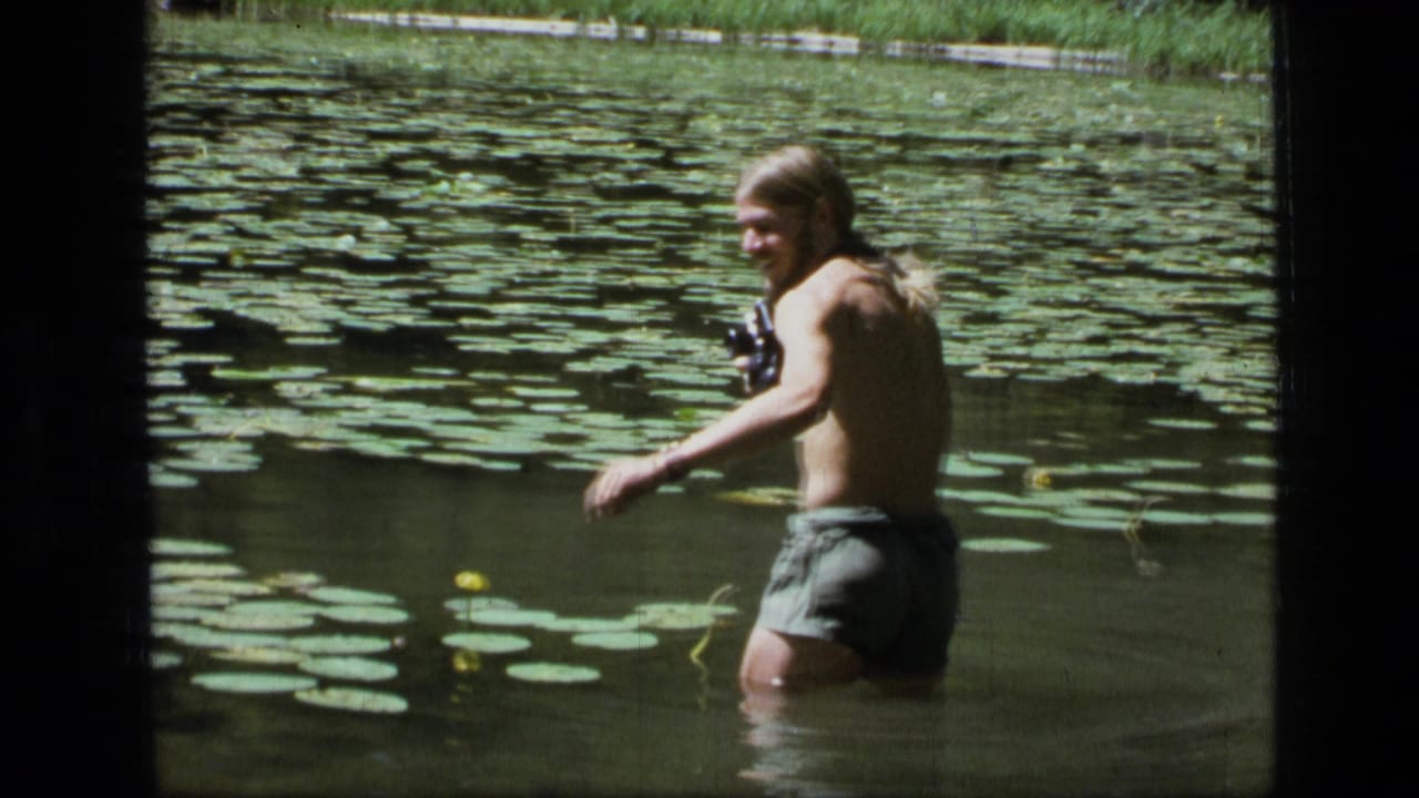 Shirtless man standing waist-deep in a lily pad pond with a camera in hand.