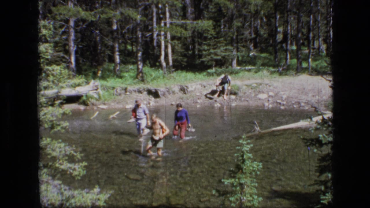 Four adults cross a clear stream in Scapegoat Wilderness, Montana, in 1973, surrounded by trees, rocks, and fallen timber.