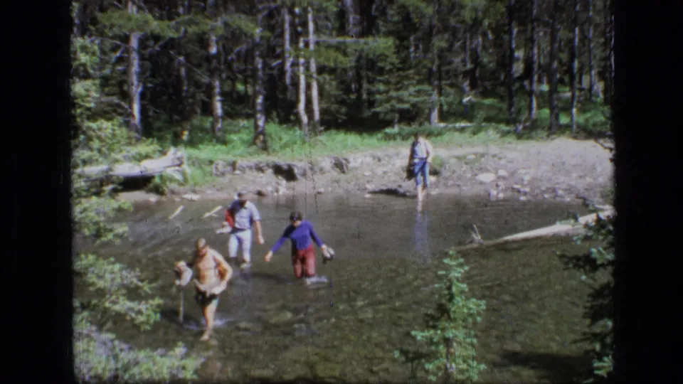 Adults wade through a clear stream in Scapegoat Wilderness, with one figure farther back near the rocky bank and dense trees beyond.