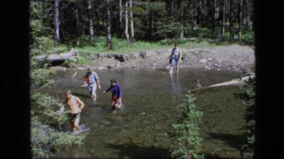 In this 1973 Montana wilderness frame, four adults wade through shallow water amid forest trees, rocks, and a fallen log.