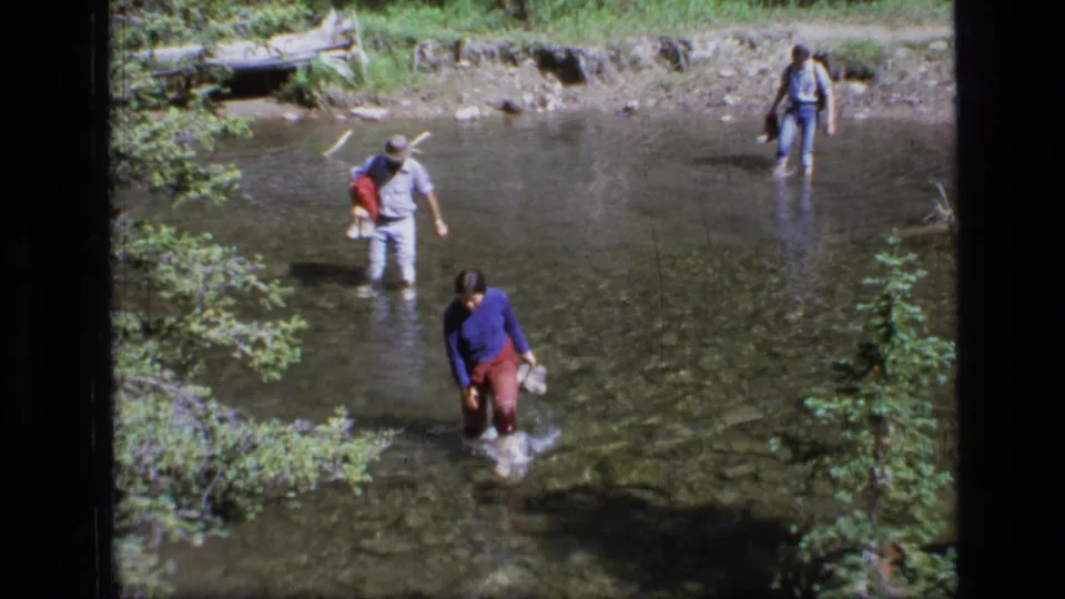 In 1973 Scapegoat Wilderness footage, several people wade through clear spring water, with one adult carrying a child behind a woman.