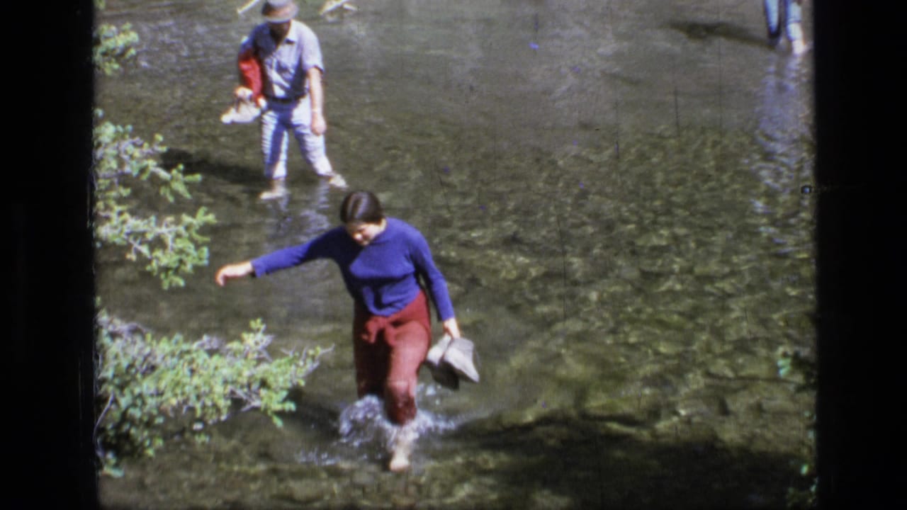 A woman in a blue sweater and red pants steps through a clear Montana stream as two other people follow in the background.