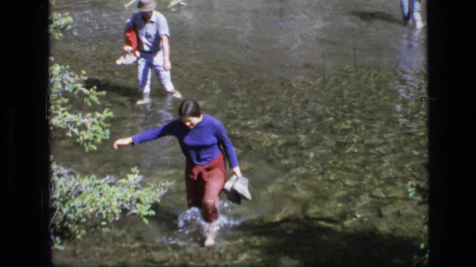 A woman in a blue sweater and red pants steps through a clear Montana stream as two other people follow in the background.