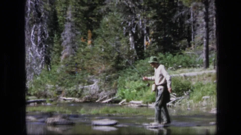A man in a green hat and light shirt stands by the water with a fishing rod in 1973 Scapegoat Wilderness home-movie footage.