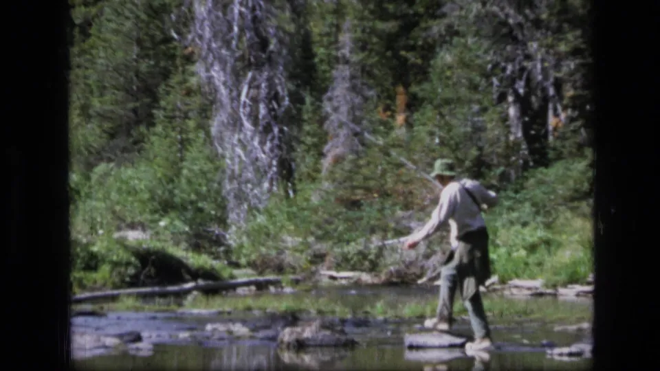 A fisherman in a hat casts beside a shallow stream with dense Montana forest visible behind him.