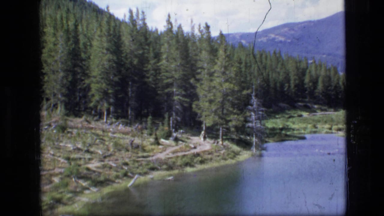 A pine-lined Montana lake and trail appear in soft 8mm footage, with mountains beyond the forested shoreline.