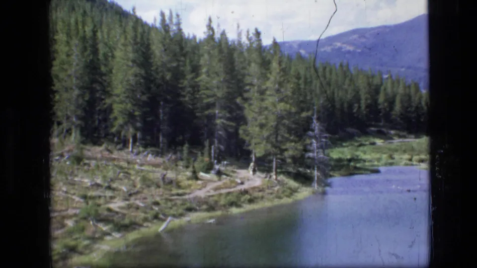 A pine-lined Montana lake and trail appear in soft 8mm footage, with mountains beyond the forested shoreline.