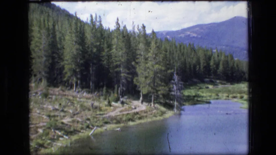 A distant hiker walks along a dirt trail beside a lake in Scapegoat Wilderness, surrounded by pines and mountain terrain.