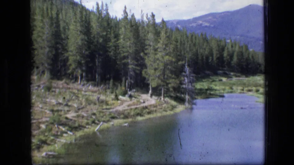 1973 8mm view of Scapegoat Wilderness, Montana, showing a forest pond, winding shoreline path, evergreens, and distant mountains.