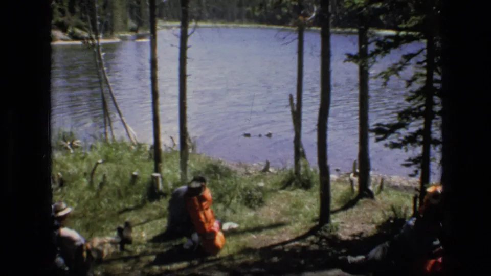 Backpackers pause by a wooded Montana lakeshore, with packs on the grass and lake water visible through the trees.