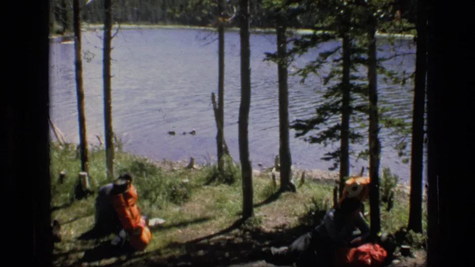 Two hikers remain seated near large packs beside a lake in archival 8mm footage from the Montana wilderness setting.