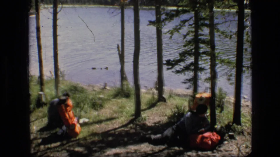 1973 Montana wilderness home-movie frame showing two distant hikers with orange packs resting near water among tall pine trunks.