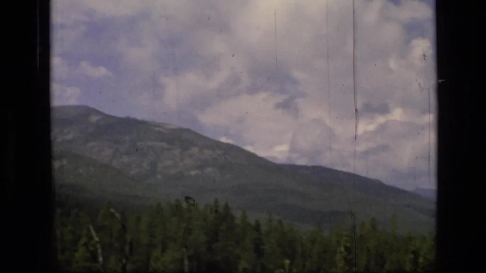 1973 Montana wilderness landscape with evergreen foreground, layered mountain ridge, and cloudy blue sky.