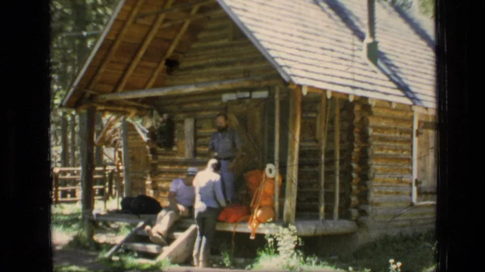 A wider 1973 Montana cabin view shows men around the steps, a sloped roof, railings, trees, and outdoor gear.