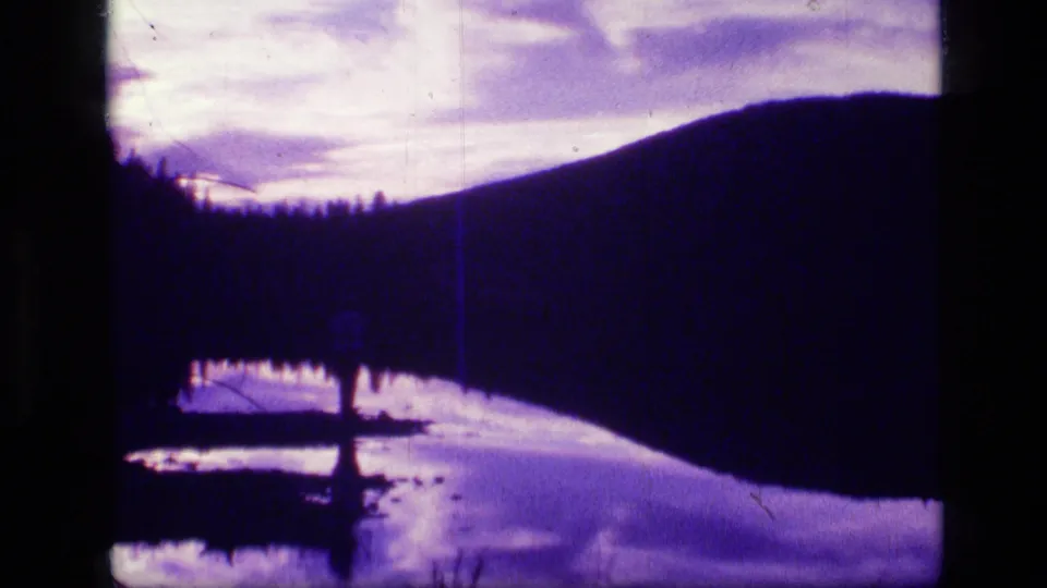 8mm Montana lake frame with dark hill, purple water reflection, and a small silhouetted shoreline figure or tree form.