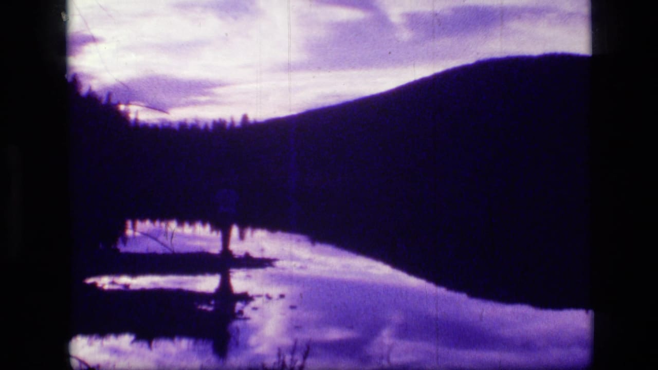 1973 8mm view of Scapegoat Wilderness, Montana, with a silhouetted ridge, tree line, reflective water, and purple-tinted sky.