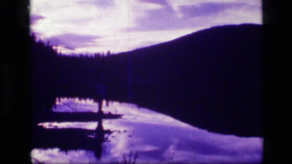 1973 8mm view of Scapegoat Wilderness, Montana, with a silhouetted ridge, tree line, reflective water, and purple-tinted sky.