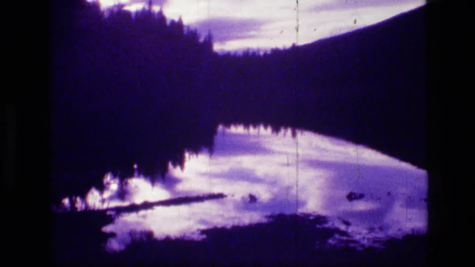 Scapegoat Wilderness landscape on 8mm film, showing a dark wooded slope, water reflection, pale clouds, and shoreline silhouettes.
