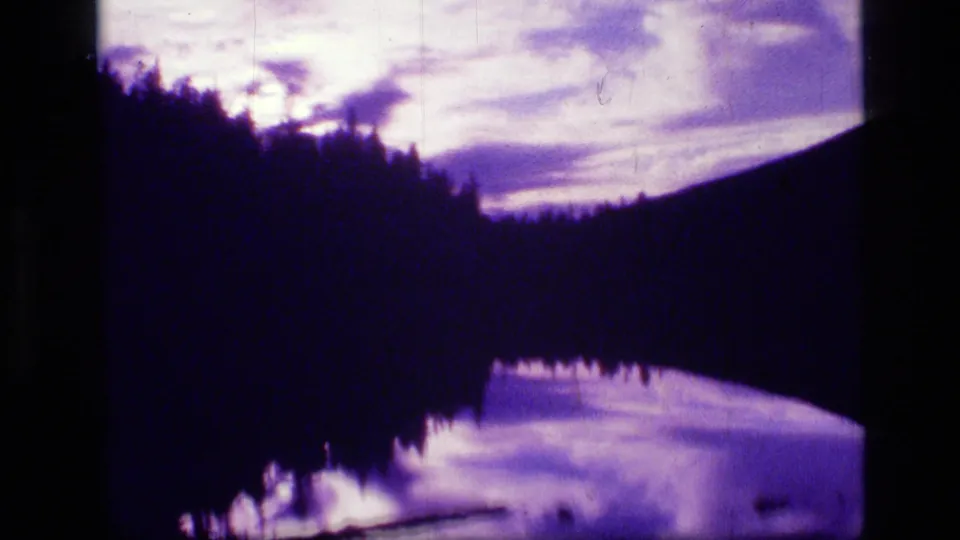 1973 Montana wilderness frame with silhouetted trees, a mountain slope, reflective water, and a small person near the shoreline.