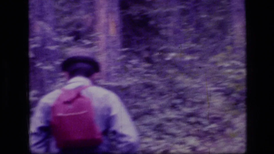 1973 home-movie frame of a man hiking from behind in Montana forest, wearing a brimmed hat and red backpack.