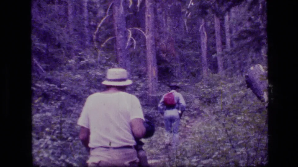 Two hikers, one in a pale hat and one with a red backpack, walk away through Scapegoat Wilderness forest in 1973 home-movie footage.