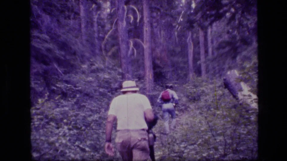 1973 8mm home-movie frame of two hikers moving along a shaded trail in Scapegoat Wilderness, Montana.