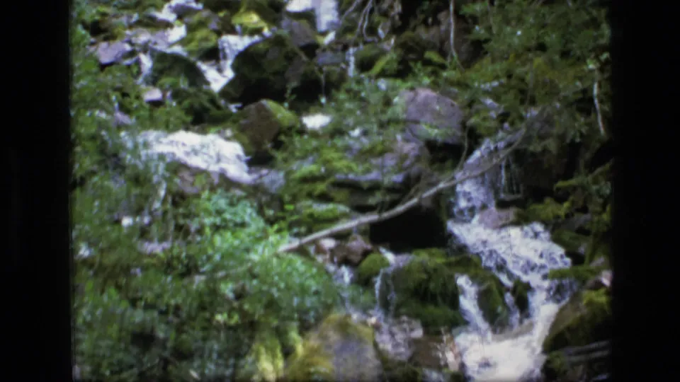 A 1973 8mm frame shows small cascades crossing mossy rocks in a wooded Montana wilderness creek.