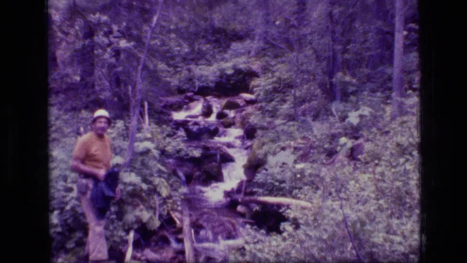 1973 Montana wilderness frame showing an adult man near a cascading forest stream, with rocks, logs, and dense trees behind him.