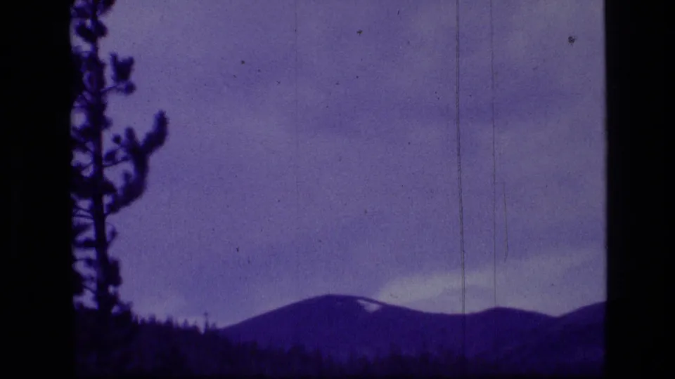Final 1973 Montana wilderness frame with a partial pine silhouette, rounded ridge, small pale snow patch, and blue-purple 8mm film color.