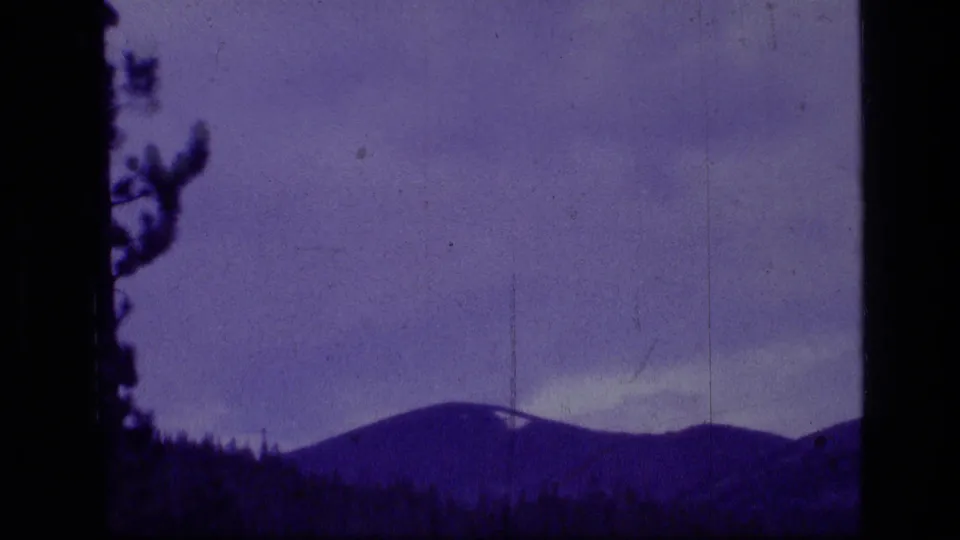 1973 8mm view of a distant mountain ridge in Scapegoat Wilderness, seen through dark pine trees with purple color shift.