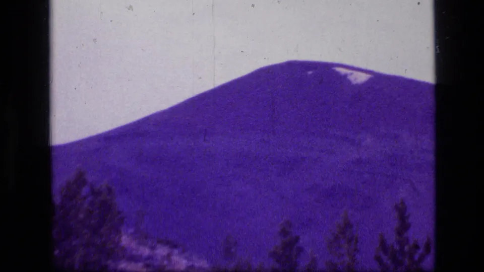1973 8mm view of a rounded Scapegoat Wilderness peak with a small snow patch and treetops below.