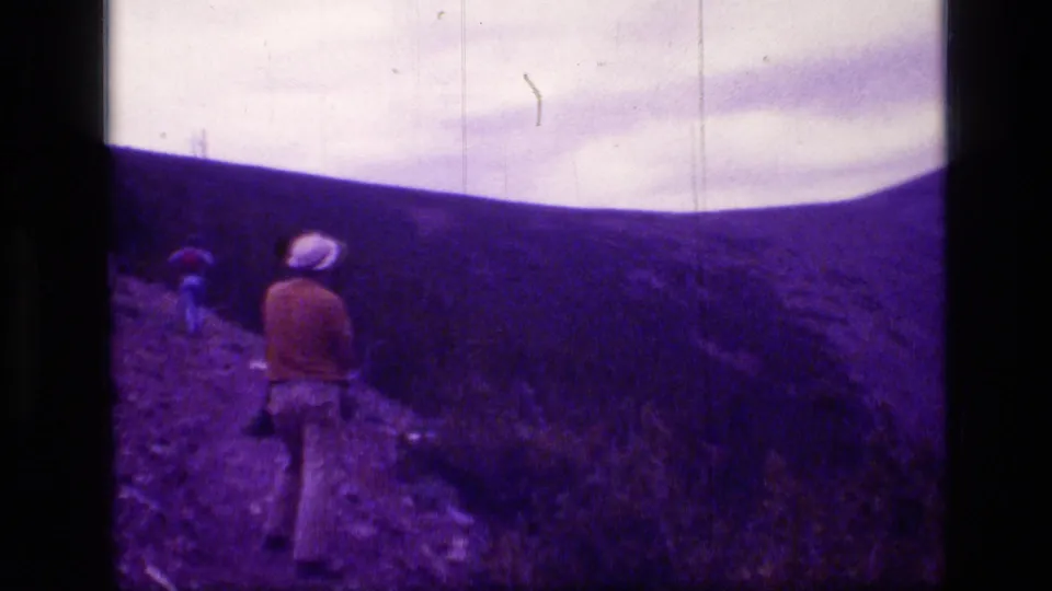 A distant hiker in casual clothing moves through a dark hillside in 1973 Scapegoat Wilderness footage.