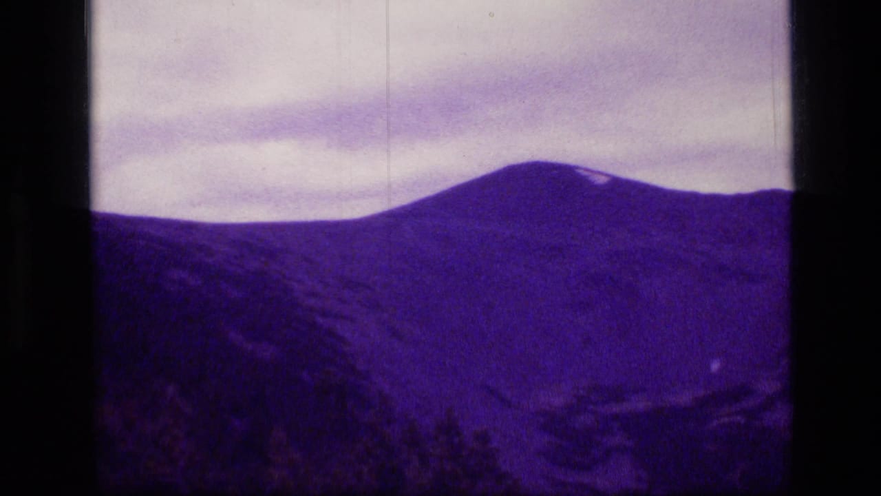 1973 8mm Scapegoat Wilderness view of a broad purple mountain ridge under pale clouds, framed by dark film borders.