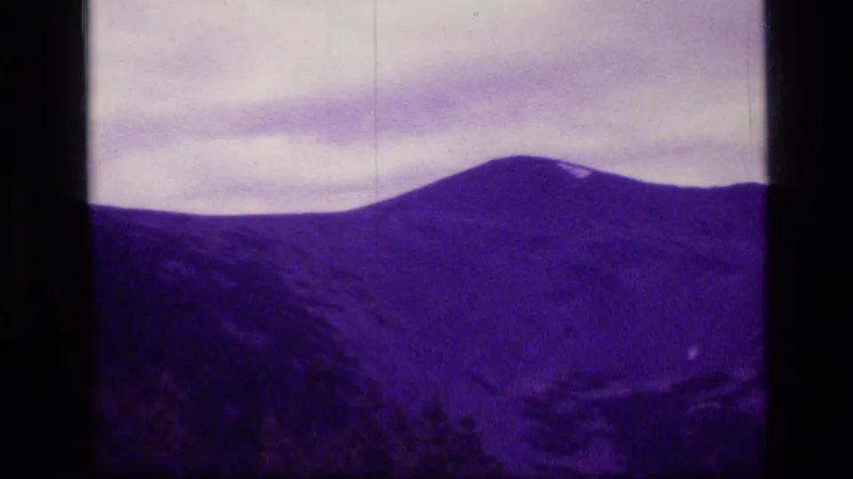 1973 8mm Scapegoat Wilderness view of a broad purple mountain ridge under pale clouds, framed by dark film borders.