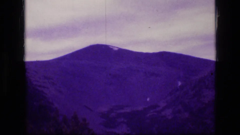 1973 Montana wilderness footage shows a wide mountain slope and cloudy sky, with grain, scratches, and strong purple dye shift.