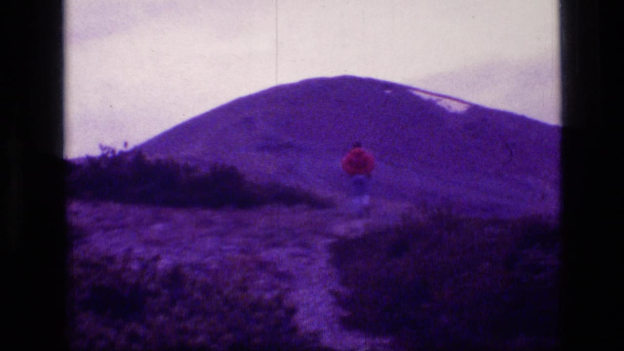 A red-jacketed hiker walks along a narrow trail in Scapegoat Wilderness, Montana, on purple-tinted 1973 8mm footage.