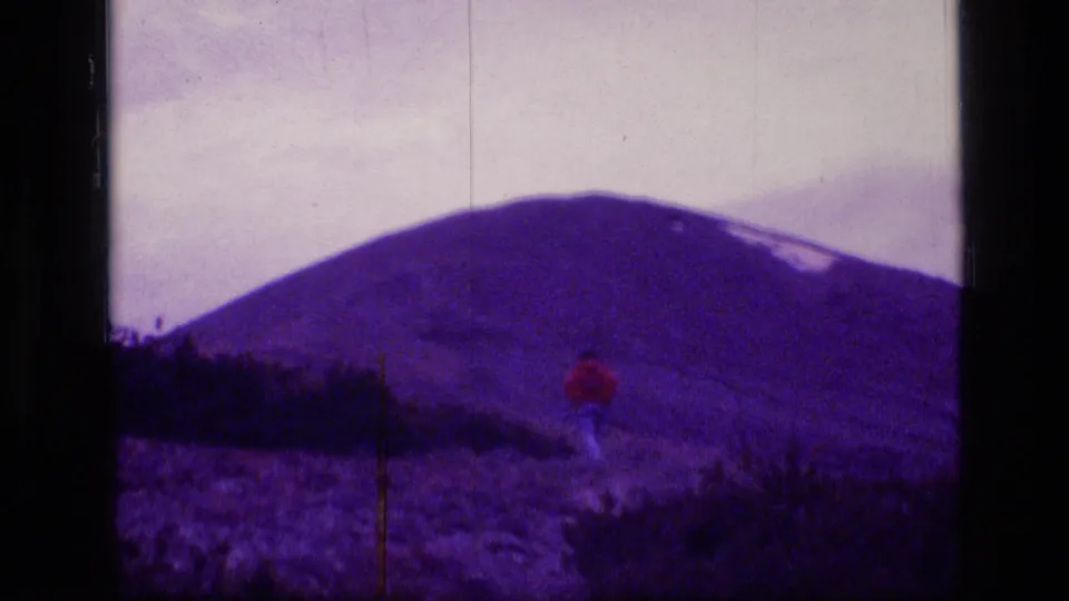 A lone hiker in red climbs through low vegetation below a broad Montana mountain ridge in archival 8mm home-movie footage.
