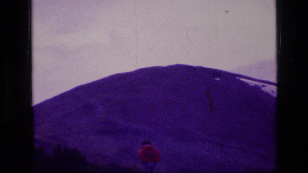 1973 8mm Montana wilderness frame with a distant red-clad walker crossing open ground below a snow-streaked mountain.