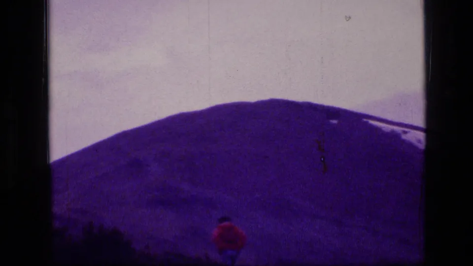 1973 8mm Montana wilderness frame with a distant red-clad walker crossing open ground below a snow-streaked mountain.