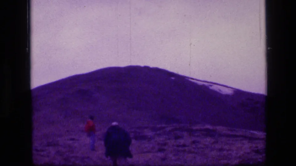1973 8mm home-movie view of two distant walkers crossing open Montana wilderness terrain below a purple-tinted mountain.