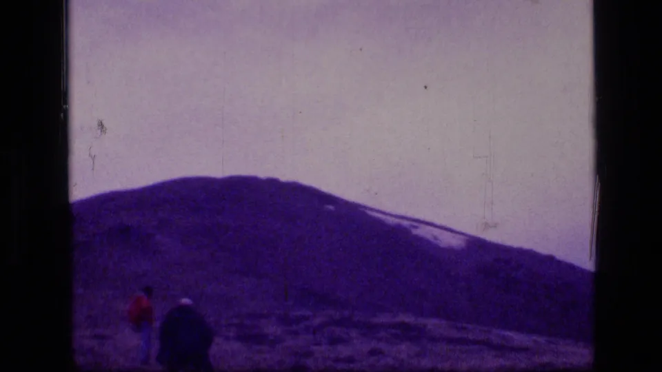 Distant figure in red and a dark pack-like shape remain in the Montana wilderness landscape beneath the snow-streaked mountain.