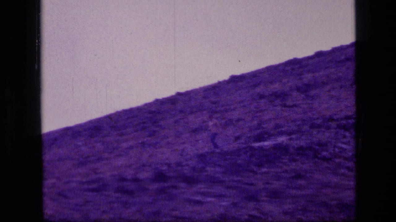 1973 8mm Scapegoat Wilderness frame with a small figure moving across a purple-tinted rocky slope beneath a pale sky.