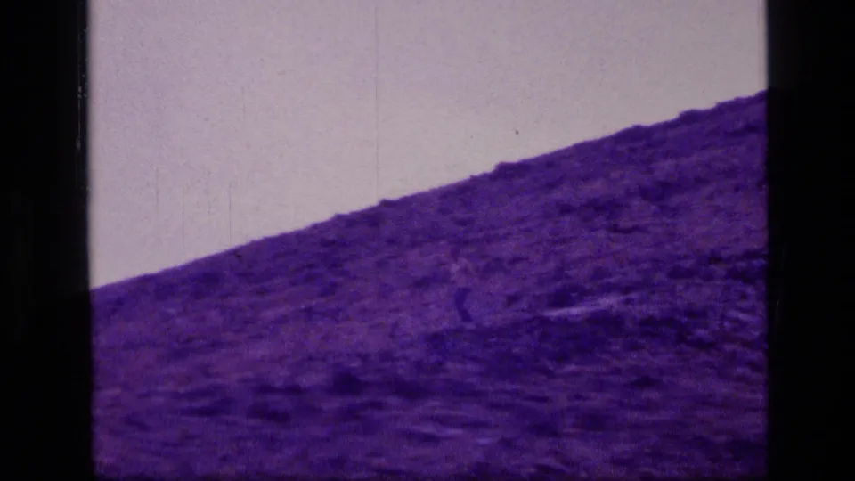 1973 8mm Scapegoat Wilderness frame with a small figure moving across a purple-tinted rocky slope beneath a pale sky.