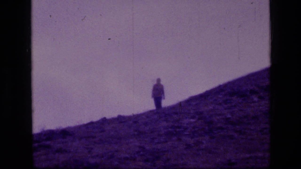 A distant person stands on a rocky mountain slope under a pale sky.